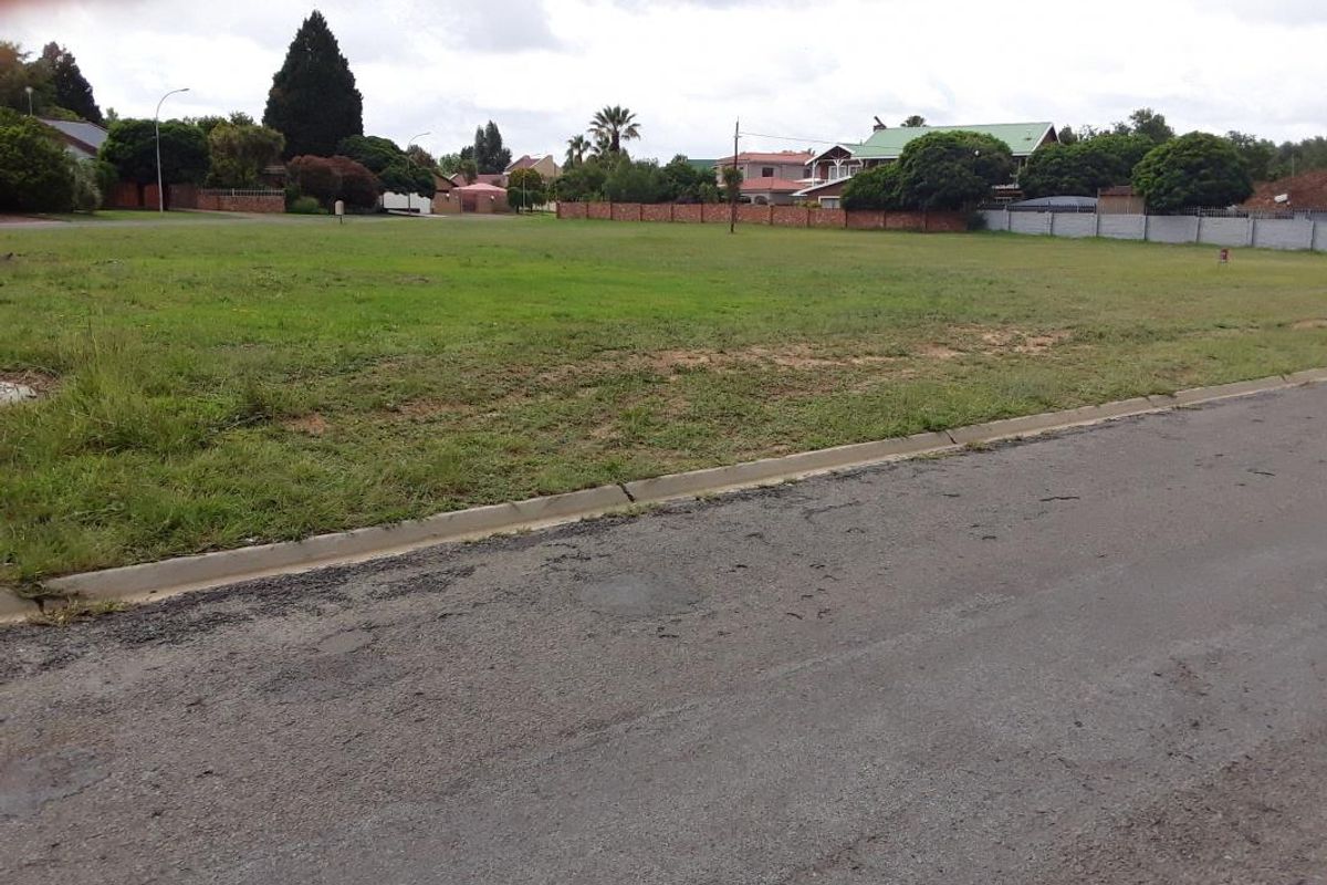 A view of a vacant residential plot from the adjacent paved street, featuring established grass and perimeter boundary walls in a suburban setting.