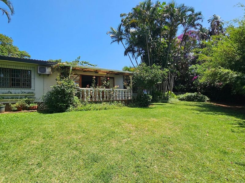 Rear view of the house featuring a covered patio with a balustrade, set within a spacious garden with mature trees and lawn.