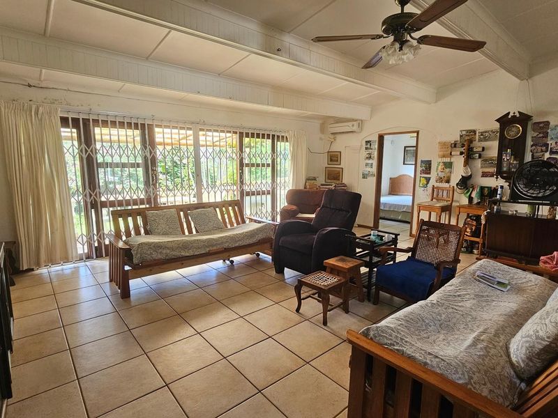 Spacious living room featuring tiled flooring, exposed beam ceilings, a ceiling fan, and large sliding glass doors with security gates leading to the patio.
