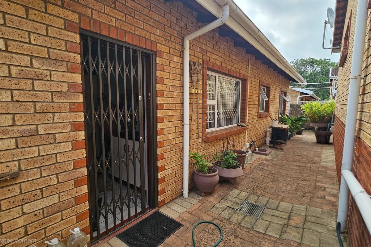 A side exterior view of a brick townhouse featuring a security gate entrance, paved walkway, and guttering system.
