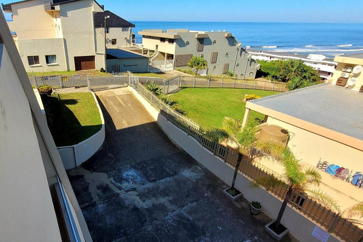 Elevated view overlooking the complex driveway, landscaped garden areas, and surrounding residential buildings with a clear ocean backdrop.