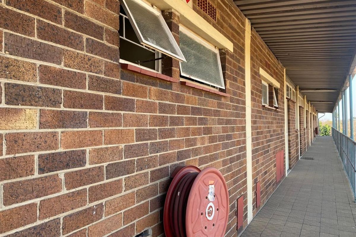 An exterior view of an apartment building walkway featuring exposed brick walls, top-hung windows, and a fire hose reel.
