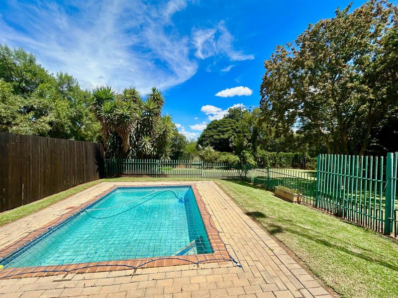 Outdoor pool area featuring a paved surround, brick coping, and secure metal perimeter fencing set within a landscaped garden.