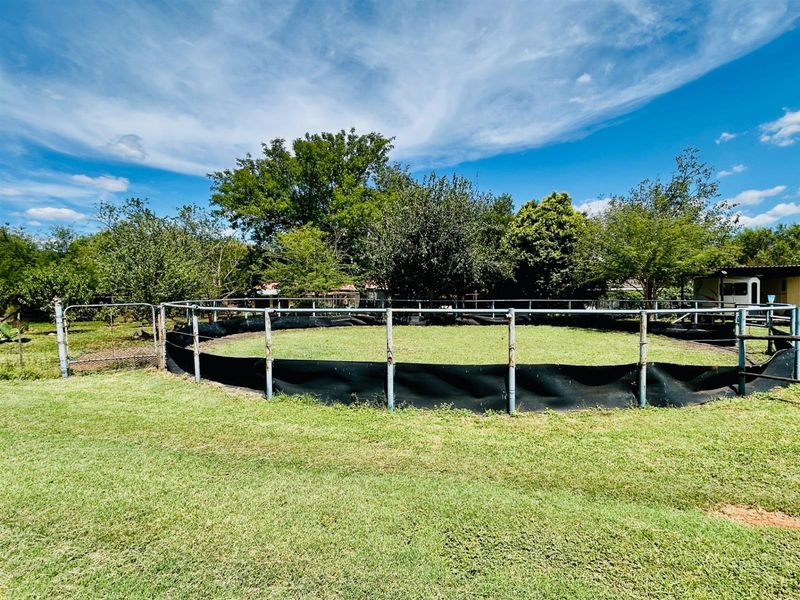 A circular outdoor training pen or enclosure featuring metal fencing and a grass surface, situated within a garden setting.