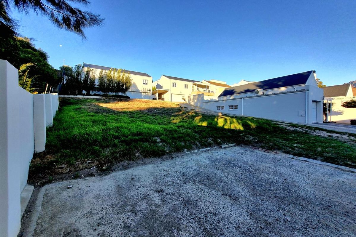 A vacant residential plot featuring a concrete driveway, boundary wall, and a gentle upward slope, situated in a developed neighborhood.