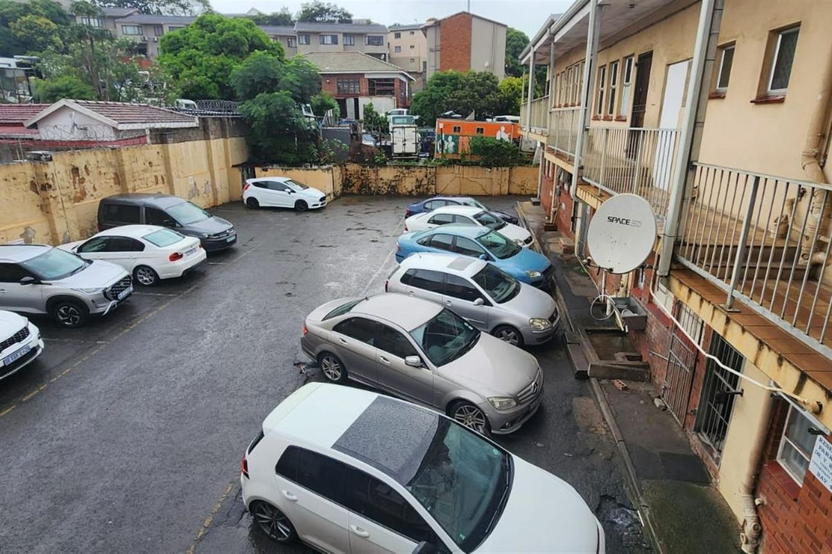 An elevated view of a retail property parking area featuring a paved lot, perimeter boundary walls, and access to the building's upper level walkway.