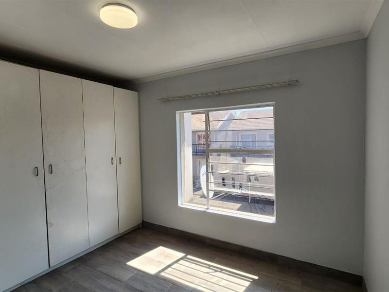 A bright, empty bedroom featuring built-in wardrobes, tiled flooring, and a window with security bars overlooking the complex.