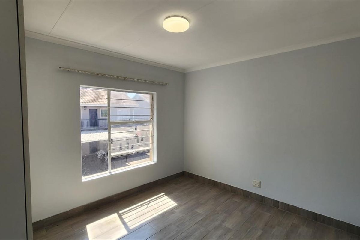 A bright, empty bedroom featuring wood-look tiled flooring, neutral walls, and a large window with security bars.