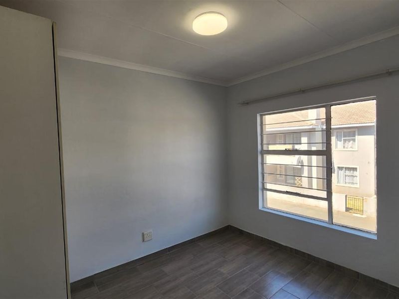 A bright, empty bedroom featuring tiled flooring, a built-in cupboard, and a window with security bars overlooking the street.