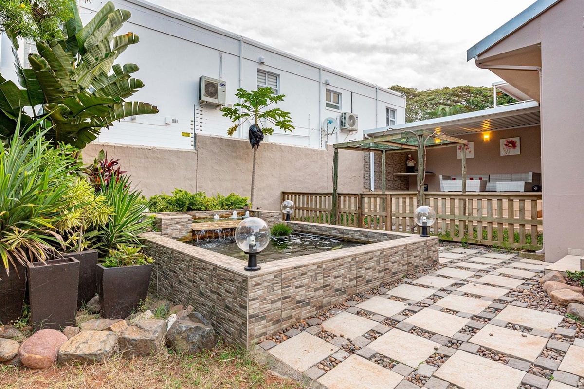 A landscaped rear garden featuring a raised stone-clad water feature, paved pathways, and a covered patio area with a wooden pergola structure.