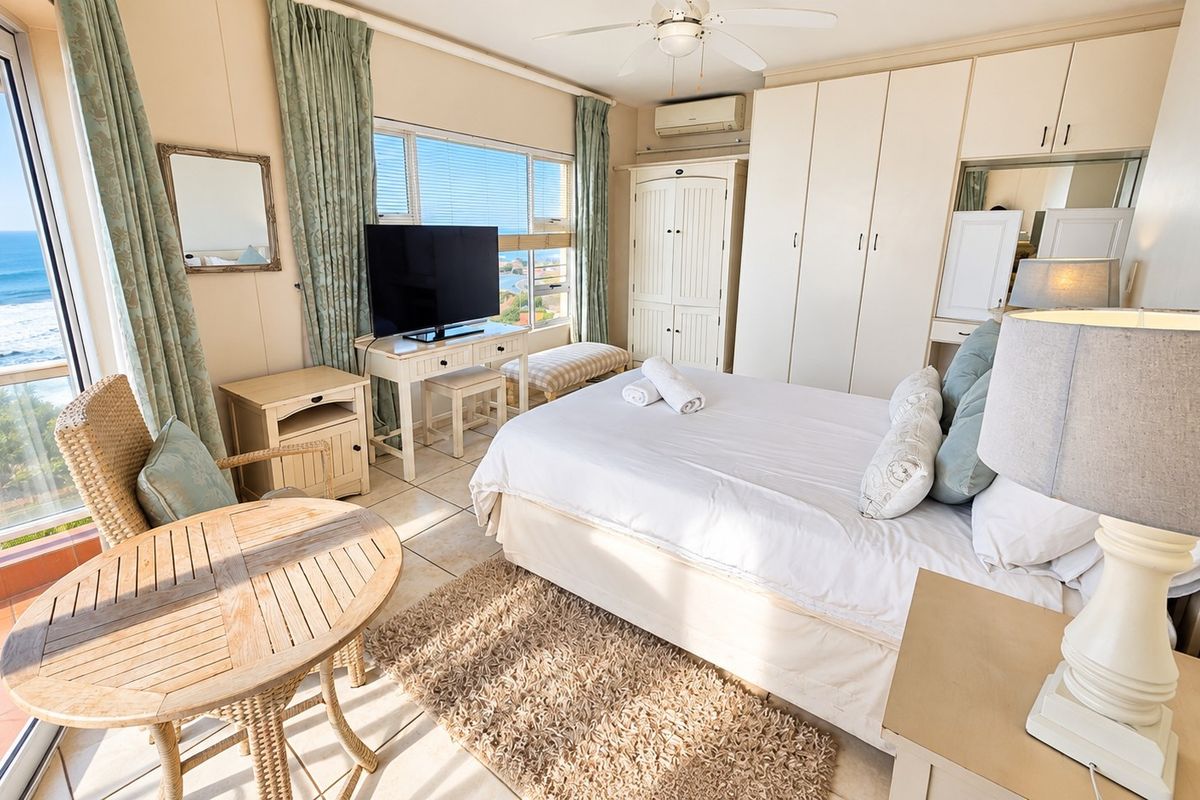 A bright bedroom featuring built-in cupboards, air conditioning, a ceiling fan, and large windows with ocean views and access to a balcony.