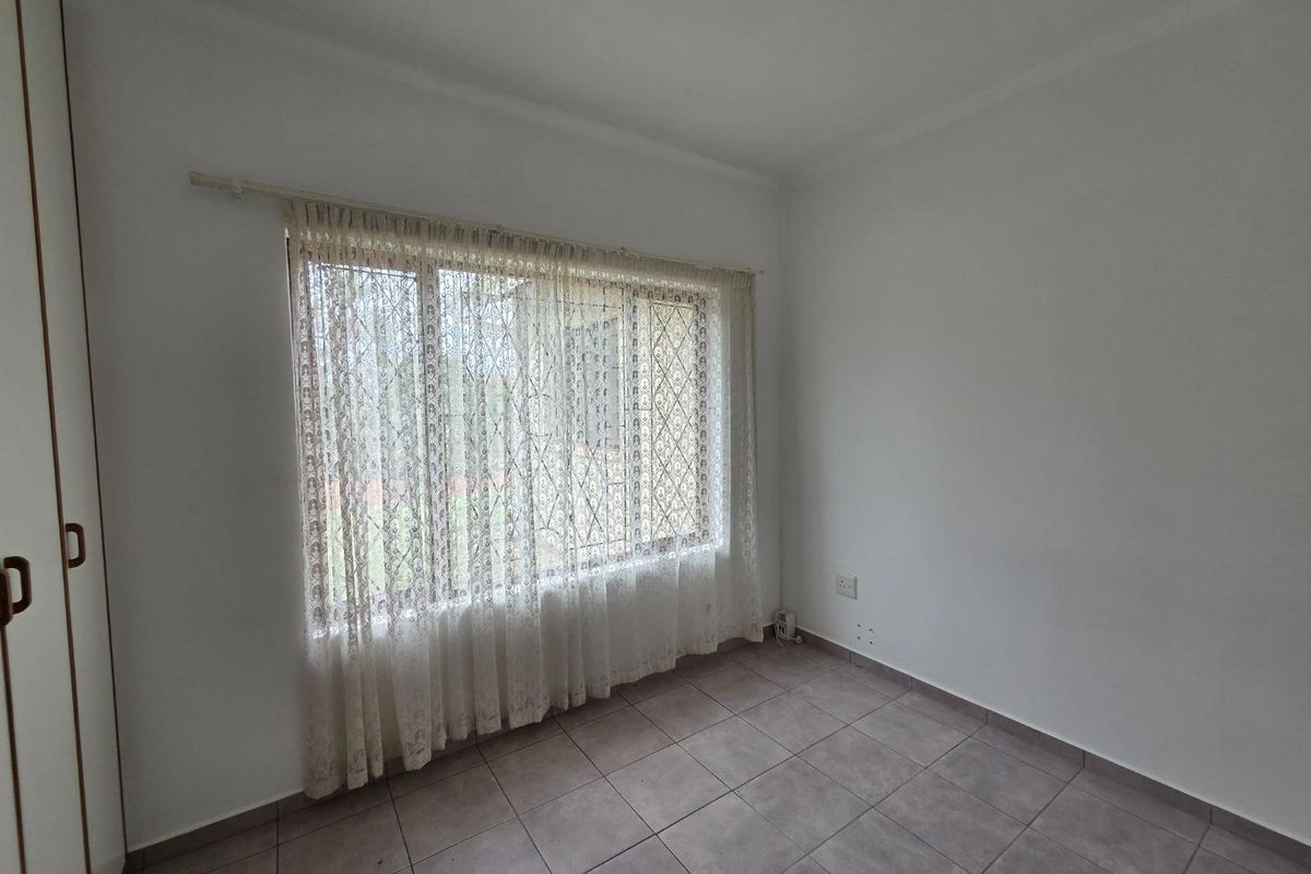 A bright, empty bedroom featuring neutral floor tiles, built-in cupboards, and a large window with curtaining.