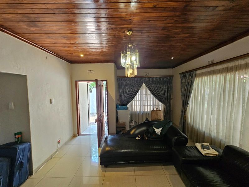 Living room featuring a distinctive dark wood-paneled ceiling, tiled flooring, and large windows with curtains, providing access to an exterior entrance.