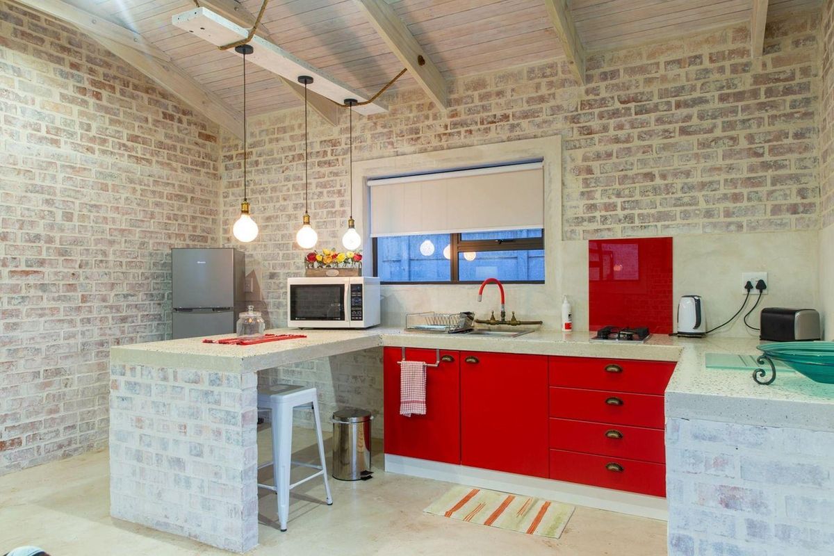A rustic-style kitchen featuring exposed brick walls, timber ceiling beams, vibrant red cabinetry, and durable stone-look concrete countertops with an integrated breakfast bar.