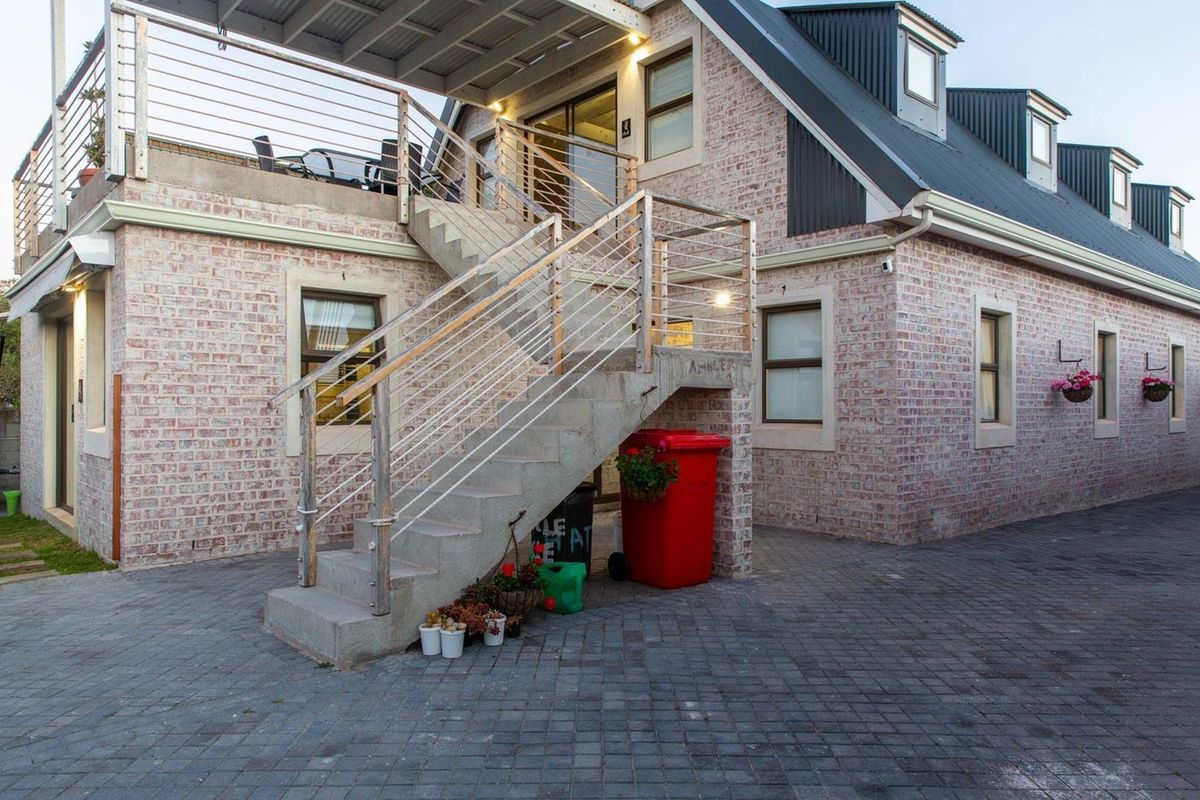 Rear exterior view of a brick house featuring an external concrete staircase leading to an upper-level balcony with metal railings and paved ground area.