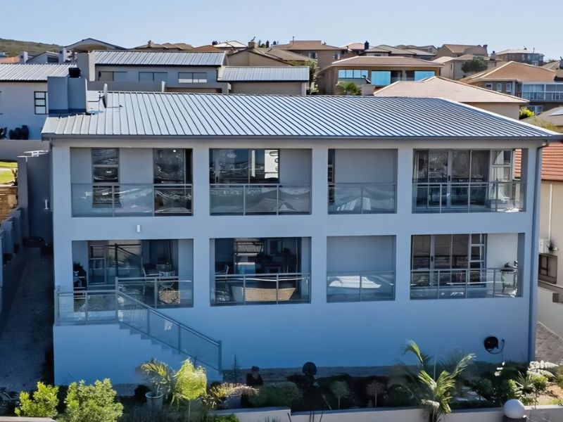A modern multi-story residential property featuring a metal roof, expansive glass balustrades, and multiple balconies overlooking a landscaped garden area.