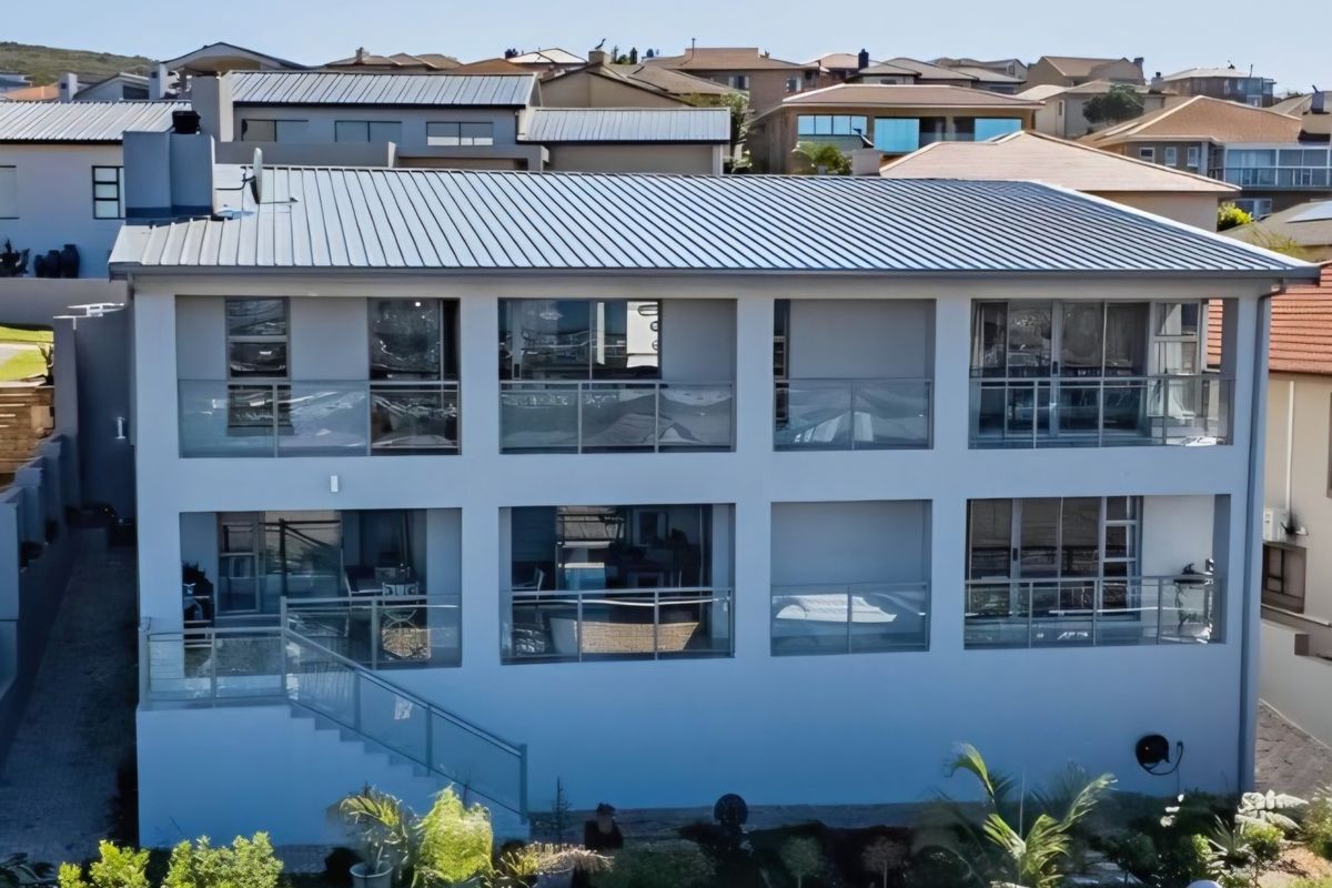 A modern multi-story residential property featuring a metal roof, expansive glass balustrades, and multiple balconies overlooking a landscaped garden area.