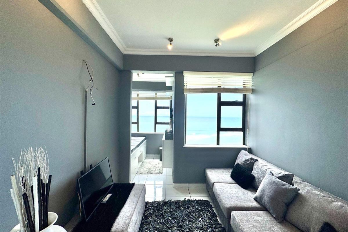 A living room featuring grey walls, tiled flooring, and large windows with ocean views, leading into an adjacent sunroom or enclosed balcony area.
