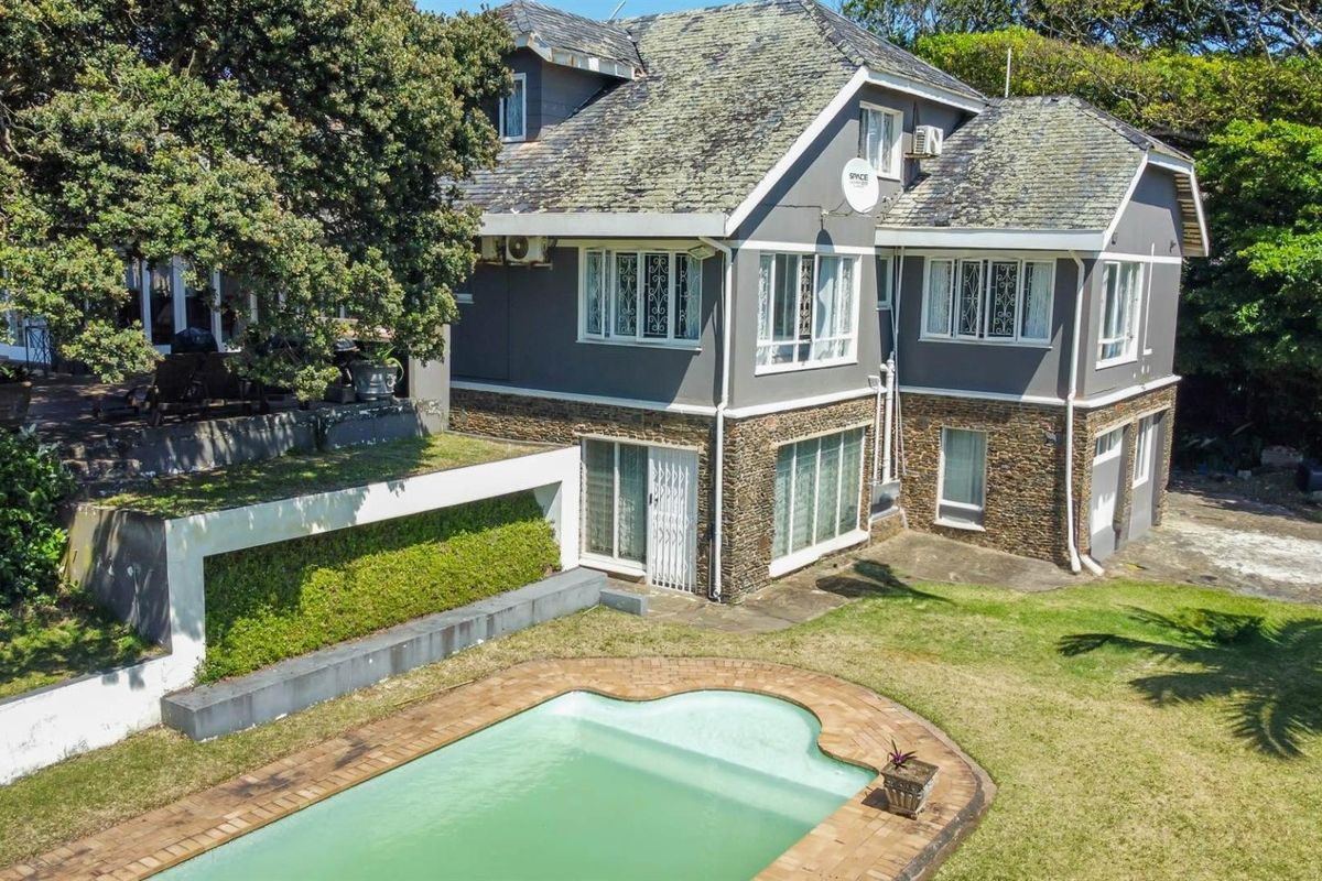 Aerial view of a multi-story residential property featuring a swimming pool, stone-clad lower facade, and mature garden surroundings.