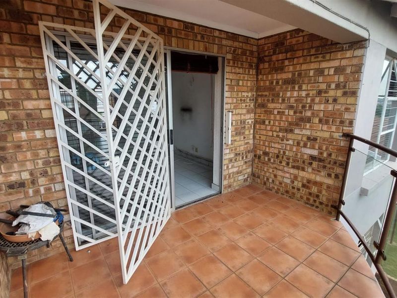 A covered balcony featuring brick walls, tiled flooring, a security gate, and a glass balustrade, offering an outdoor space for relaxation.