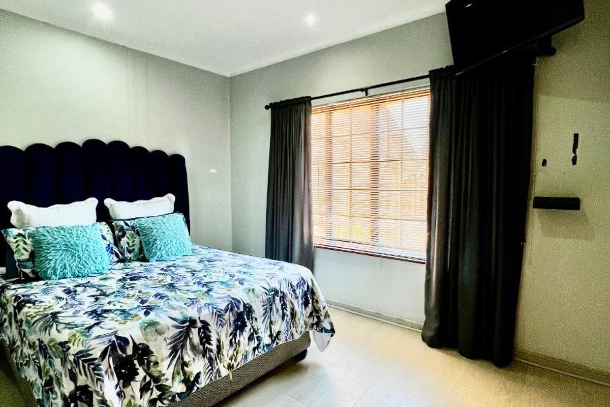 A well-maintained bedroom featuring light-toned laminate flooring, recessed downlighting, and window treatments including venetian blinds and curtains.