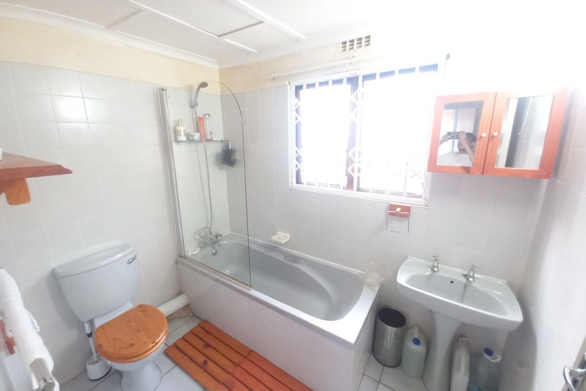 A functional bathroom featuring a bathtub with shower screen, pedestal basin, and toilet, finished with white wall tiles and security window bars.