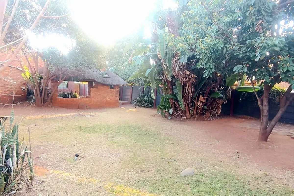 A spacious rear garden featuring a thatched lapa structure, mature trees, and perimeter fencing, offering a private outdoor area.