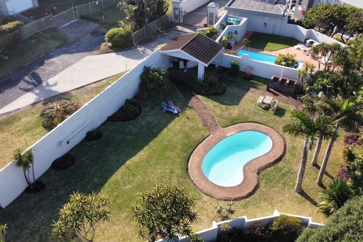 Aerial view of a spacious backyard featuring a kidney-shaped swimming pool, a covered patio area, a built-in braai, and established garden landscaping.