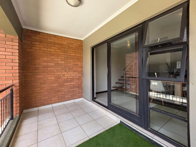 A covered balcony featuring tiled flooring, a feature brick wall, and large black-framed sliding glass doors leading into the apartment interior.