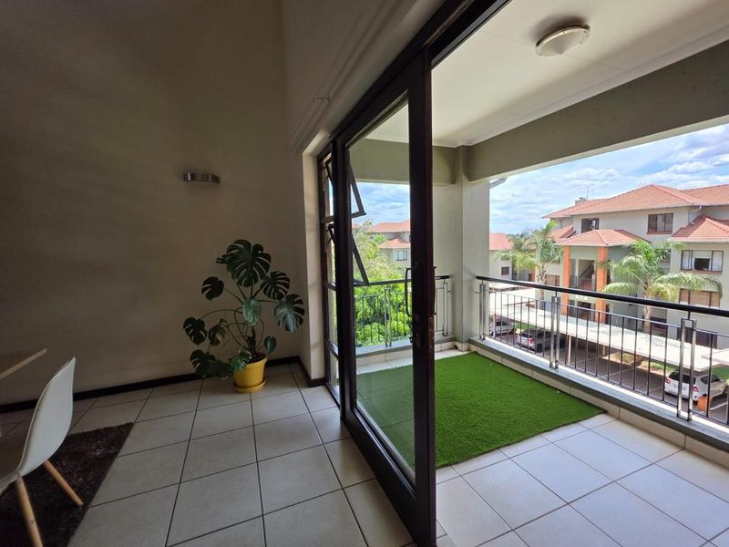 A bright indoor living area featuring large sliding glass doors that open onto a private balcony with artificial turf and metal railings.