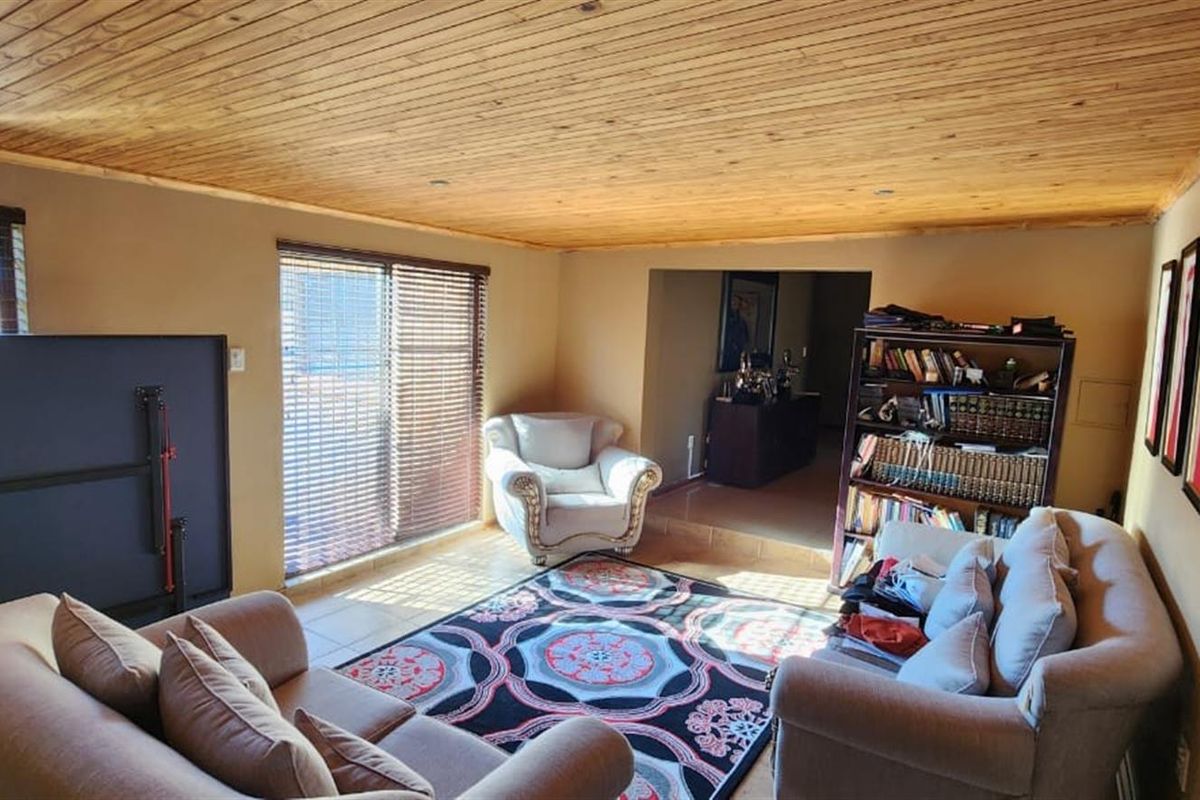 A living room featuring a distinctive timber-clad ceiling, tiled flooring, and large windows with fitted blinds, providing access to an adjacent space.