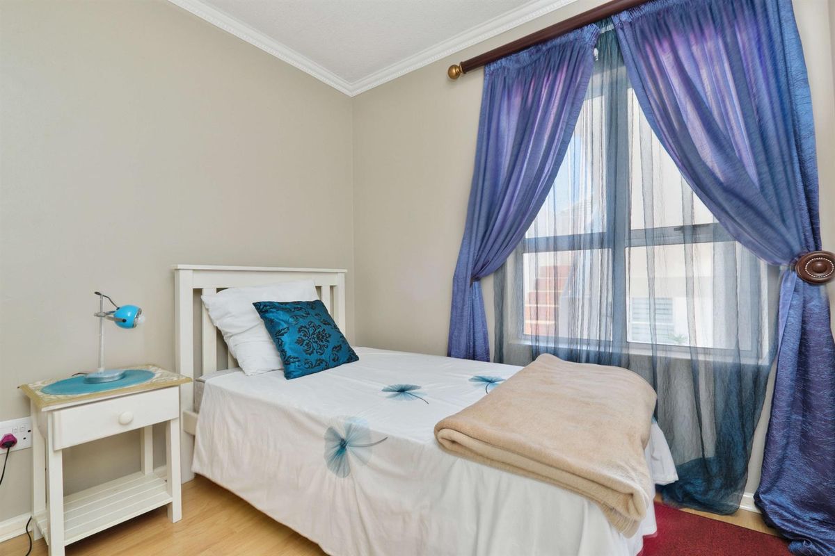 A compact bedroom featuring light-toned laminate flooring, neutral wall paint, and a window with decorative curtain treatments.