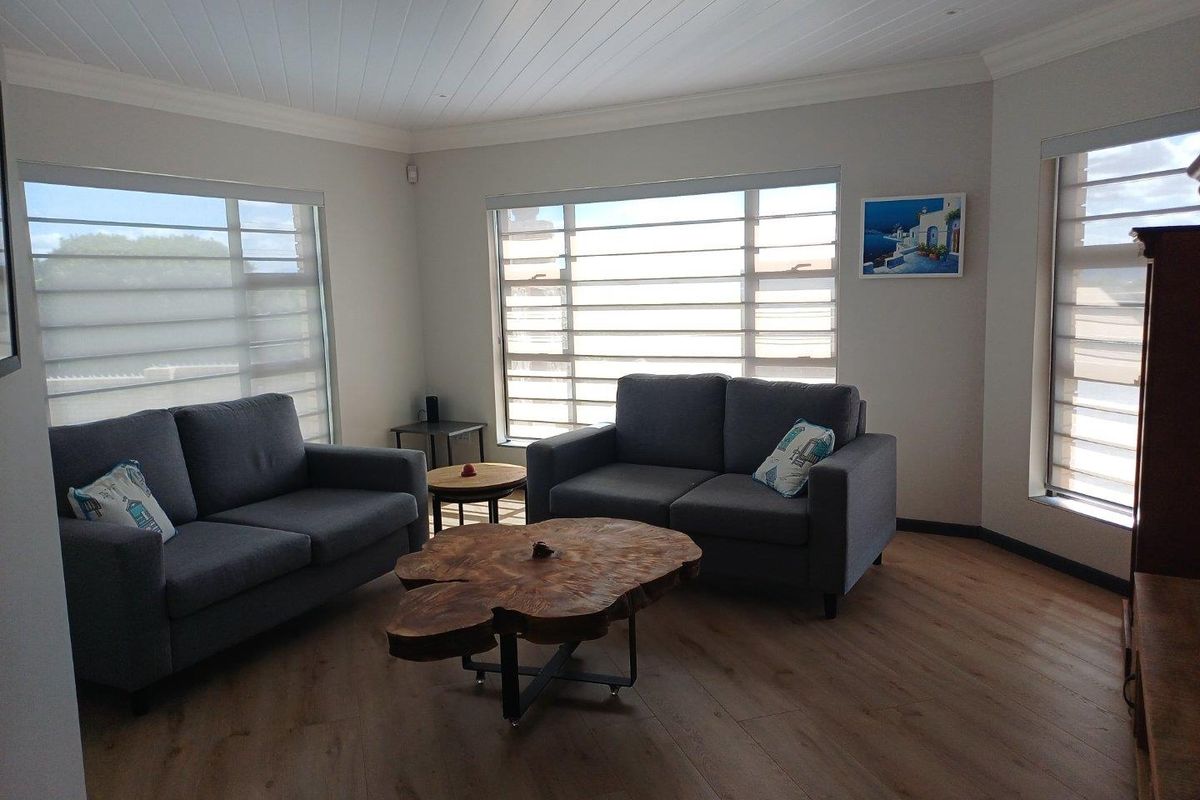 A bright and modern living room featuring wood-look flooring, large windows with fitted blinds, and a clean, neutral aesthetic.