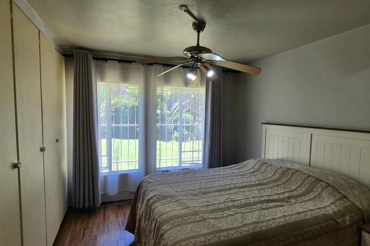 A bedroom featuring wood-look flooring, built-in cupboards, a ceiling fan, and large windows with security bars overlooking the garden.