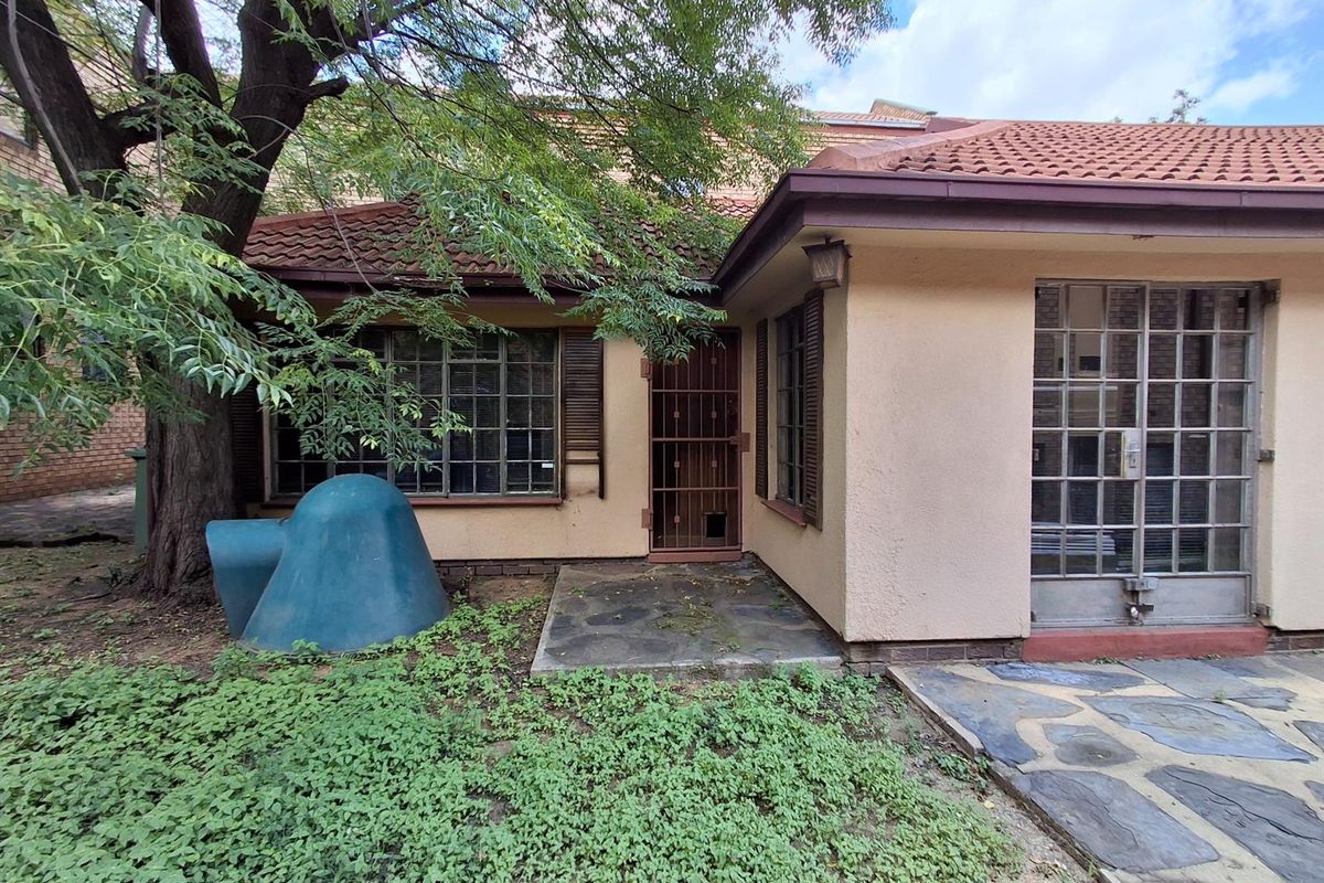 Exterior view of the office building featuring a tiled roof, security gates, and a paved patio area surrounded by mature trees and garden space.