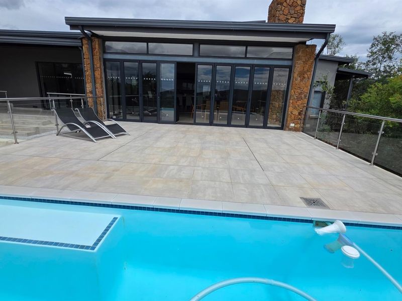 Spacious outdoor patio area featuring large format tiling, glass balustrades, and direct access to a swimming pool with stone-clad chimney feature.
