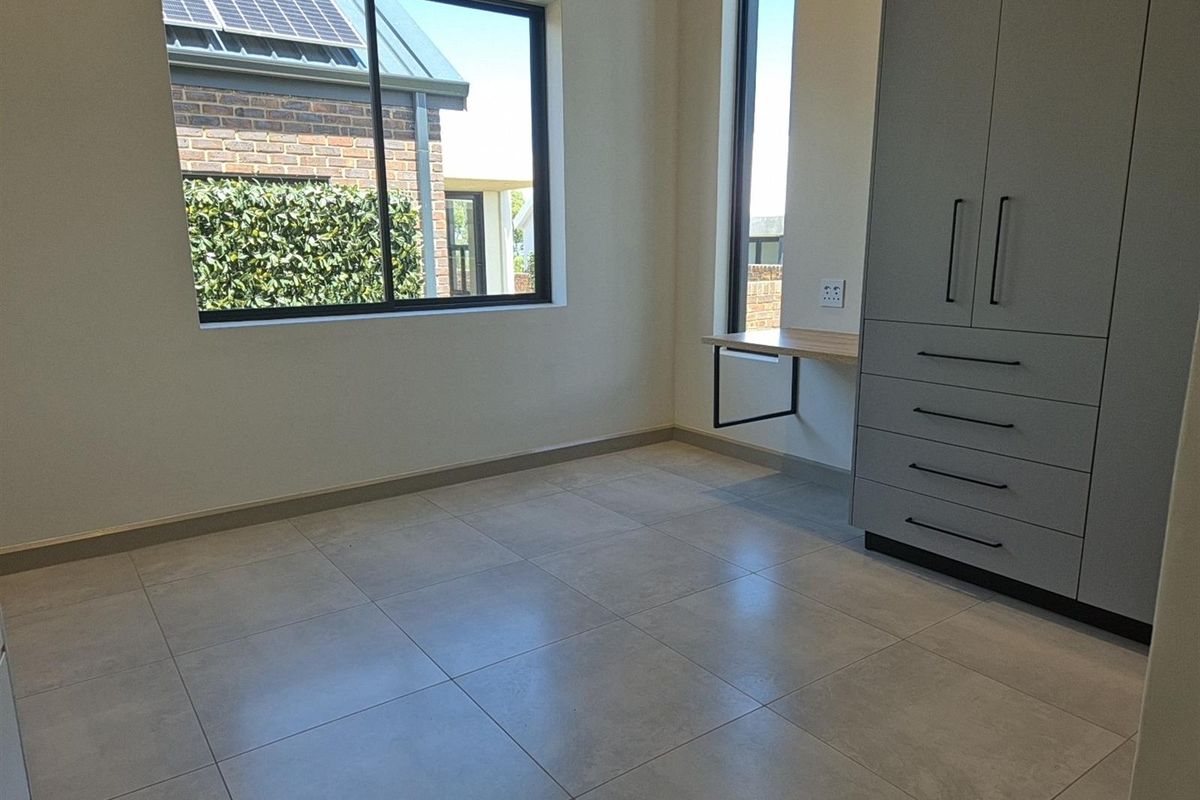 A modern, empty bedroom featuring large windows, tiled flooring, and a built-in grey wardrobe with an integrated desk space.