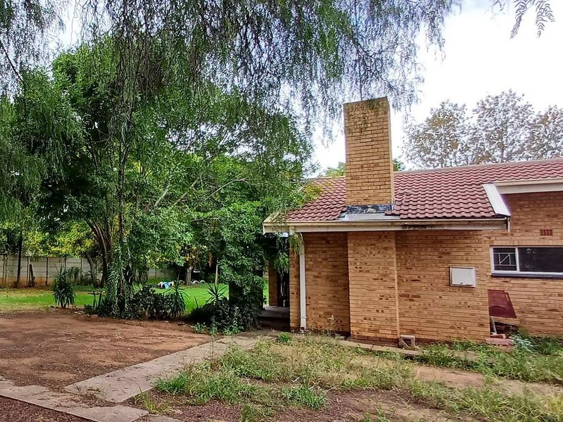 Rear view of a brick house featuring a prominent chimney, tiled roof, and an expansive, unmaintained garden area with mature trees.