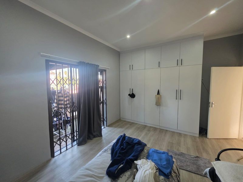 A well-maintained bedroom featuring modern built-in cupboards, light laminate flooring, recessed downlights, and a sliding door leading to an outdoor area with security gate.