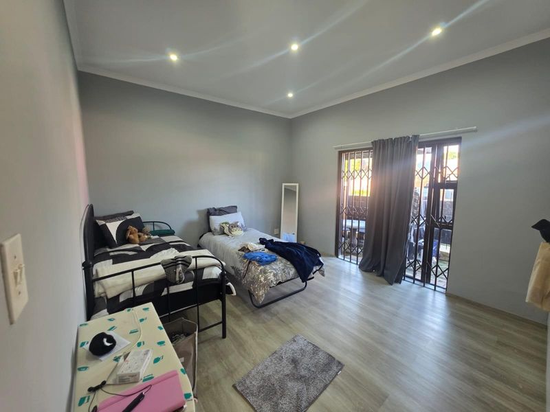 A well-lit bedroom featuring modern laminate flooring, recessed ceiling lighting, and a sliding glass door with a security gate providing access to the exterior.