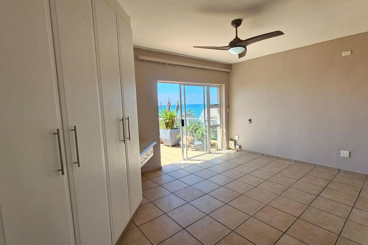 A bright bedroom featuring built-in wardrobes, tiled flooring, a ceiling fan, and sliding glass doors opening onto a balcony with ocean views.