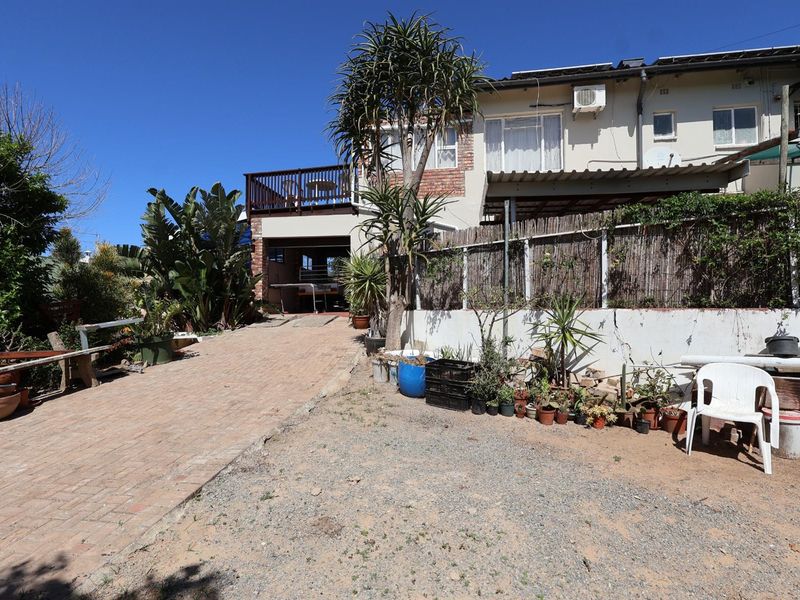 Rear view of the property featuring a paved driveway, a raised balcony, and a garden area with a retaining wall.
