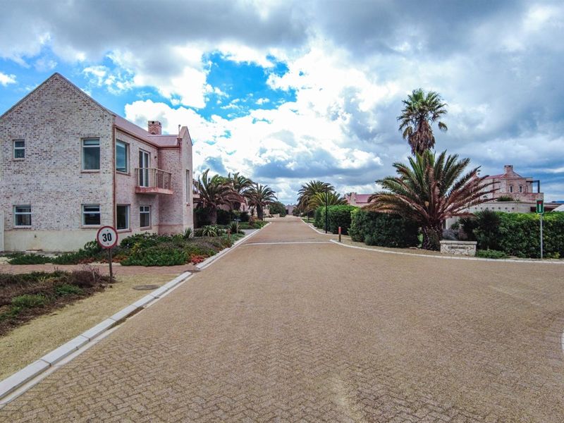 A view of a paved residential street within a well-maintained estate, featuring brick-faced buildings, landscaped gardens, and mature palm trees.