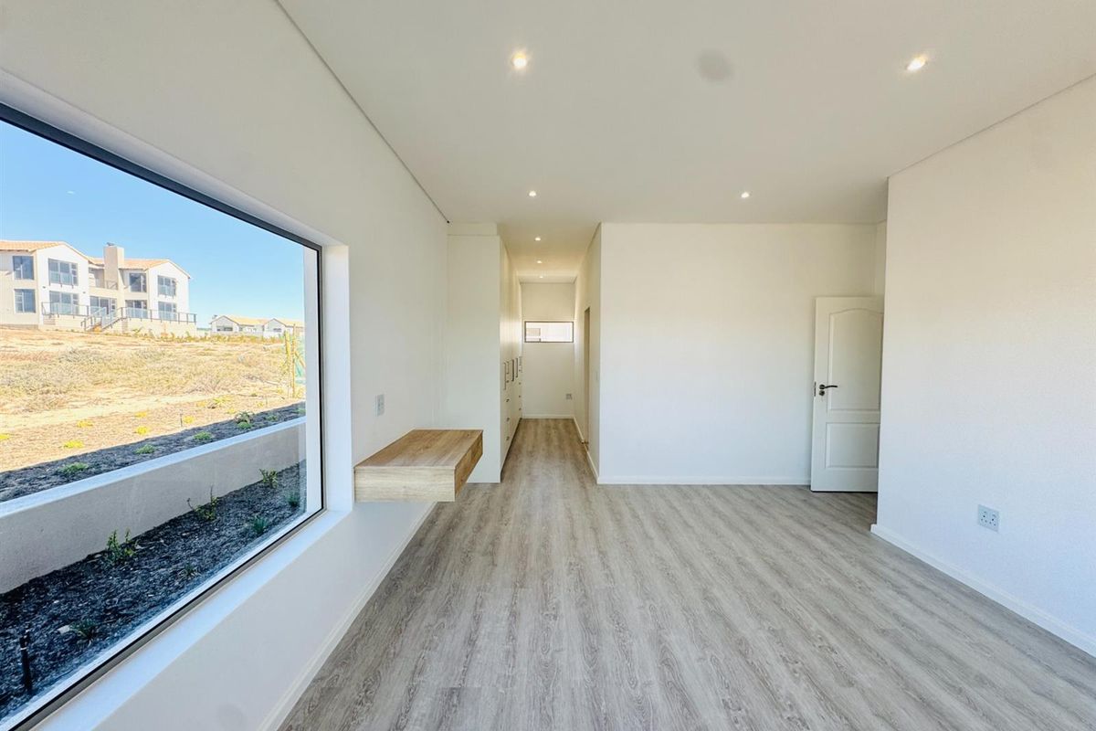 A bright, modern bedroom featuring light wood-look flooring, recessed ceiling lighting, a built-in floating desk, and floor-to-ceiling built-in cabinetry.