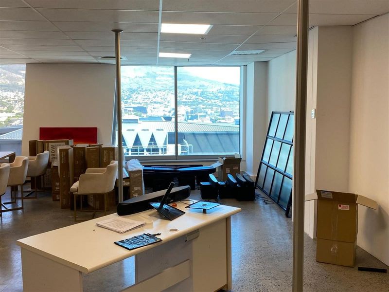 An open-plan office space featuring large floor-to-ceiling windows with city views, suspended ceiling tiles, and polished concrete flooring.