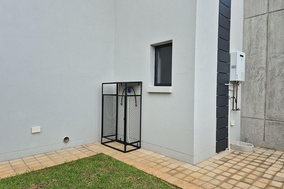 Exterior side area featuring a gas water heater, paved walkway, small lawn, and a secure metal enclosure for gas cylinders.
