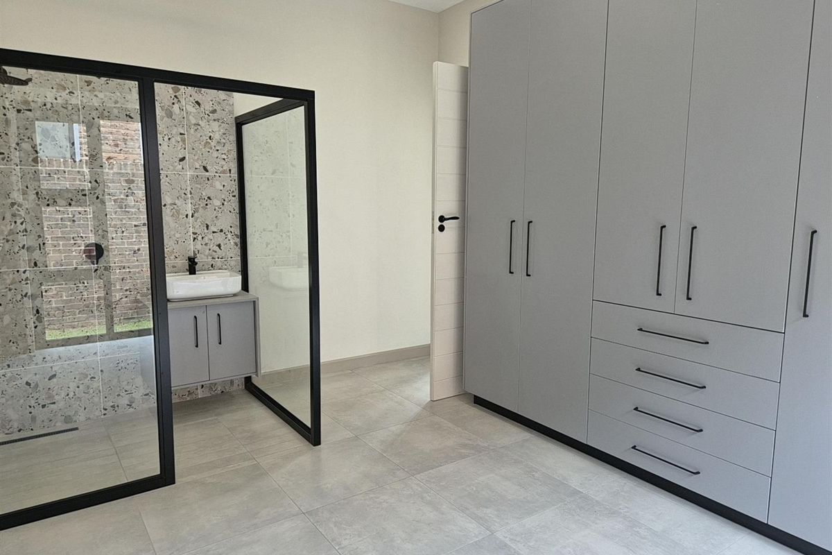 Modern bedroom featuring large built-in grey cabinetry and an open-plan en-suite bathroom with a glass-enclosed shower and terrazzo-style tiling.