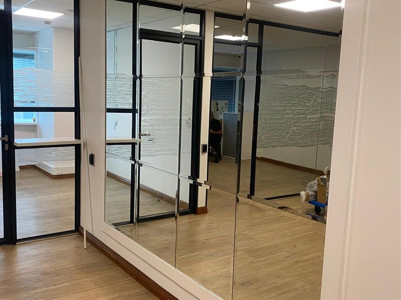 Modern office interior featuring glass-partitioned walls, wood-look flooring, and a large decorative mirrored wall panel.