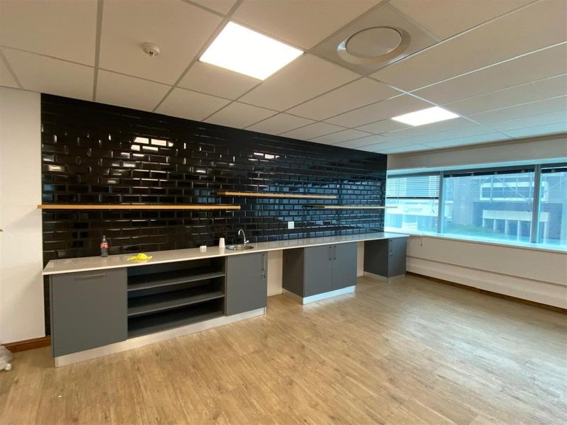 A modern office kitchenette featuring grey cabinetry, a stainless steel sink, and a striking black subway tile backsplash with floating wooden shelves.