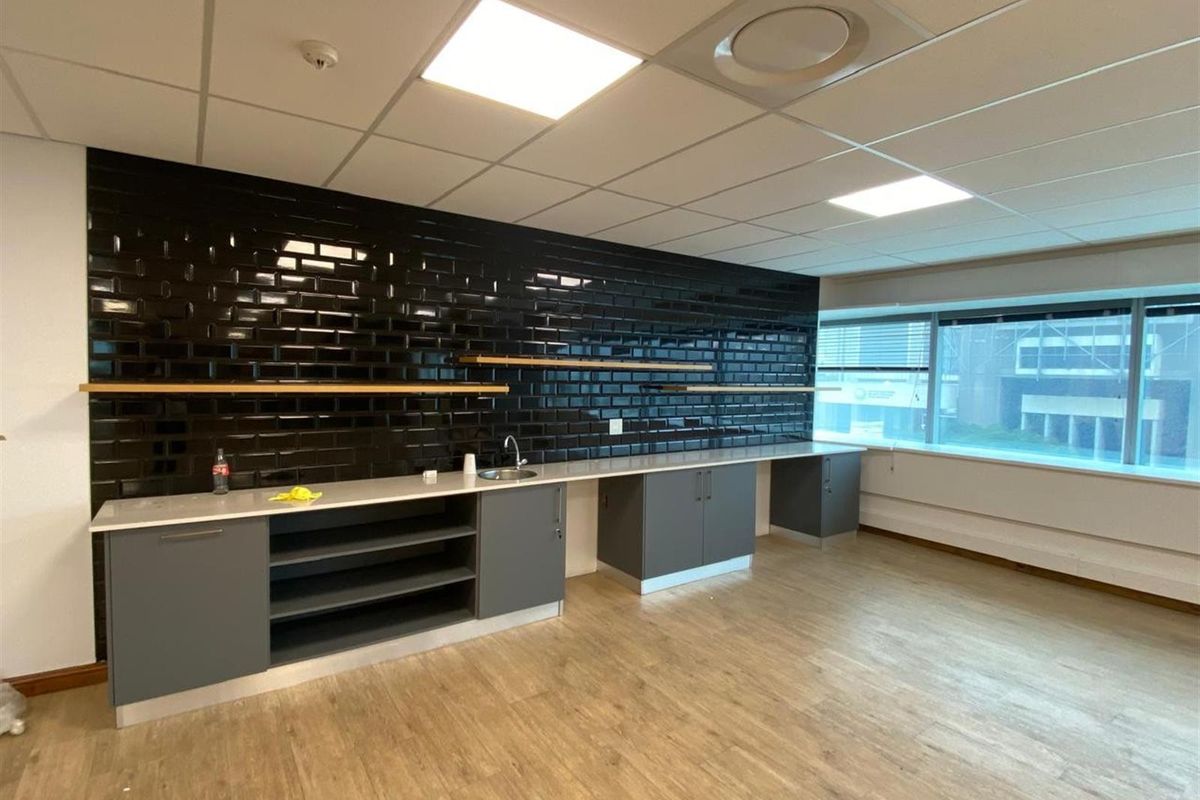 A modern office kitchenette featuring grey cabinetry, a stainless steel sink, and a striking black subway tile backsplash with floating wooden shelves.